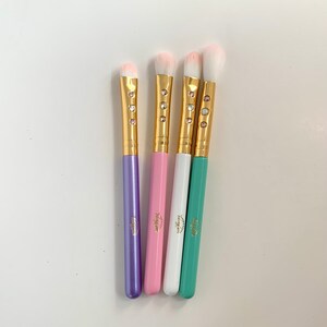 Op de afbeelding: Vier make-up kwasten met gouden handvatten en witte haren. De handvatten zijn versierd met strass steentjes en hebben de tekst "Fairy Glam" erop gedrukt. De kwasten zijn paars, roze, wit en groen.