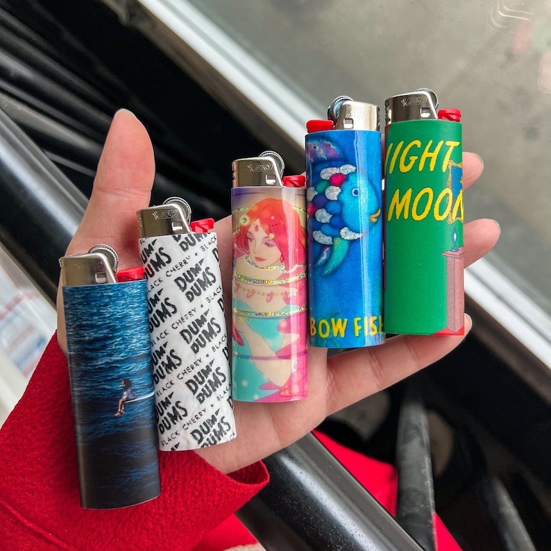 May include: A collection of five lighters with various designs. One features a person surfing on a wave, another has a woman with red hair, and a third displays a blue fish with the text "BOW FISH". The fourth lighter has the text "NIGHT MOON".