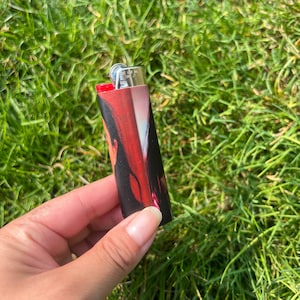 May include: A black and red lighter with a printed image of a person. The lighter has a red ignition button and a silver metal top. The lighter is held against a background of green grass.