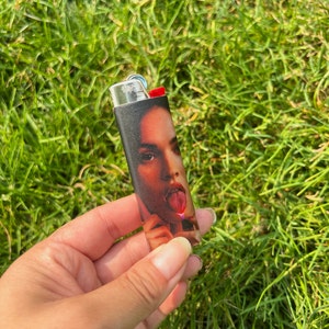 May include: Black and silver lighter with a red ignition button. The lighter features a full-color image of a woman with her tongue out. The lighter is held against a background of green grass.