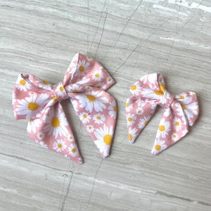 May include: Two pink fabric hair bows with a white daisy print. The larger bow is about 4 inches wide and the smaller bow is about 2 inches wide.