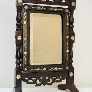 Puede incluir: Espejo de tocador de madera oscura con tallas ornamentadas e incrustaciones florales. El espejo rectangular está enmarcado con un borde oscuro y se apoya en un soporte decorativo. La parte superior del espejo tiene tallas.