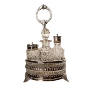 May include: An antique silver-plated vanity set with three glass bottles and a small jar. The bottles have decorative stoppers and are set on a detailed, ornate base with a handle. The set is designed for perfumes or toiletries.