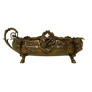 Puede incluir: Jardinera antigua de metal dorado con forma rectangular. La jardinera presenta detalladas tallas en relieve, un motivo central de instrumento musical y asas decorativas. Se apoya sobre cuatro patas en forma de pezuña, mostrando una estética vintage.