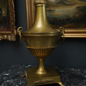 Puede incluir: Una urna dorada con tapa, asas y base de pedestal. La urna tiene un cuerpo acanalado y un adorno decorativo en la parte superior. La base es cuadrada con detalles ornamentados. El fondo incluye pinturas enmarcadas.