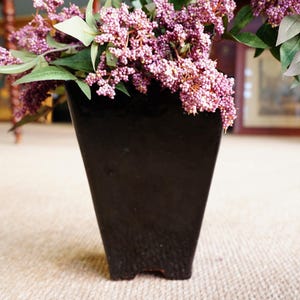 May include: A black, square-shaped vase with a smooth finish holds a dense arrangement of artificial green leaves and clusters of small, light purple berries that spill over the top edges of the container in a natural, overflowing display.