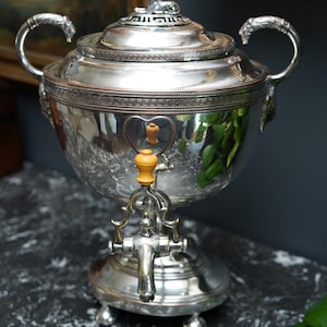Puede incluir: Una urna de té de metal plateado con cuerpo esférico, dos asas ornamentadas y un grifo con mango de madera clara. La tapa está rematada con una pequeña figura de león y el borde está decorado con un patrón de greca.