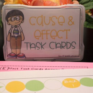 Puede incluir: Una caja de plástico transparente con una etiqueta colorida que dice "Cause & Effect Task Cards". La etiqueta presenta una ilustración de dibujos animados de un niño con gafas y una camisa a rayas. La caja está sobre una superficie rosa.