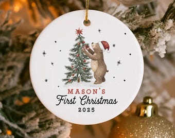 May include: Round white Christmas ornament featuring a bear decorating a Christmas tree. The ornament reads "MASON'S First Christmas 2025" and is decorated with small black stars. A gold string is attached for hanging.