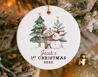 May include: A white ceramic Christmas ornament with a gold hanging loop. The ornament features a watercolour illustration of a bear and snowman wearing Santa hats, with two Christmas trees. Text reads "Jacob's 1st Christmas 2025."