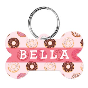 May include: Bone-shaped pet tag with a pink background and a pattern of donuts with chocolate and vanilla frosting. The name "BELLA" is printed in white block letters across the center. A silver metal ring is attached for easy attachment to a collar.