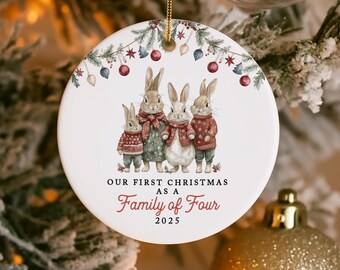 May include: A round white Christmas ornament with a family of four rabbits in festive attire. The ornament has a floral border and the text "OUR FIRST CHRISTMAS AS A Family of Four 2025". The ornament is hanging from a gold string.