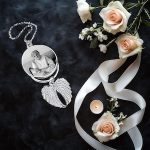 May include: A silver memorial pendant with a photo of a person, hanging from a chain. The pendant has a heart-shaped wing design. The image also includes white roses, a white ribbon, and a lit candle on a dark background.