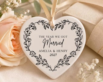 May include: A heart-shaped ceramic ornament with a floral border and the text "THE YEAR WE GOT Married AMELIA & HENRY 2027." The ornament has a cream-coloured background and a gold ribbon for hanging.