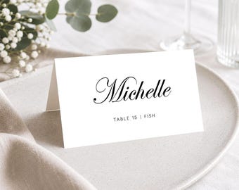 May include: A white folded place card featuring the name Michelle in a black elegant script font, with the text TABLE 15 | FISH printed in a smaller sans-serif font below. The card is positioned upright on a plate, displaying a clean design.