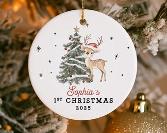 May include: A round white Christmas ornament with a gold hanging loop. It features a festive design with a Christmas tree and a deer in a Santa hat. The text reads "Sophia's 1st Christmas 2025". Small black stars are scattered around.