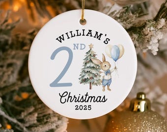 May include: Round white ceramic Christmas ornament with a gold hanging cord. The ornament has the text "William's 2nd Christmas 2025". A watercolour illustration of a bunny holding balloons and a Christmas tree is also on the ornament.
