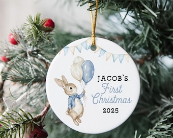 May include: A round white Christmas ornament featuring a bunny illustration holding balloons. Text reads "JACOB'S First Christmas 2025". The ornament has a gold hanging string and is decorated with blue and beige bunting.