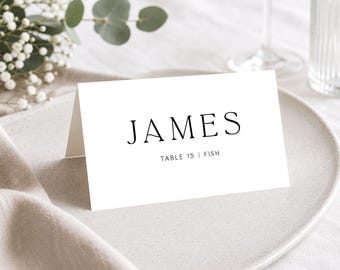 May include: A white folded place card with black text. The name JAMES is printed in a large serif font, with the text TABLE 15 | FISH printed in a smaller sans-serif font below it. The card is made of heavy cardstock and stands upright.