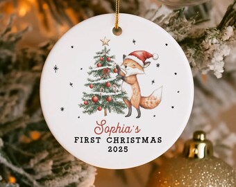 May include: A round white ceramic Christmas ornament with a fox in a Santa hat decorating a Christmas tree. The text "Sophia's FIRST CHRISTMAS 2025" is in red and black. The ornament has a gold string for hanging and is decorated with small black stars.