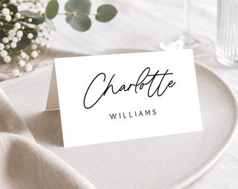 May include: A white folded place card featuring the name Charlotte in a black cursive script font, with the surname WILLIAMS printed below in a simple black sans-serif typeface. The card is positioned upright on a neutral-coloured plate.