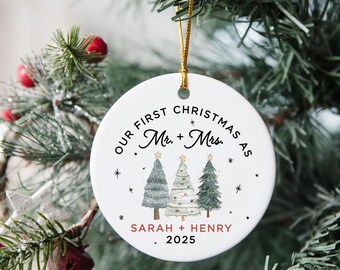 May include: A white ceramic Christmas ornament with gold string, featuring the text "Our First Christmas as Mr. + Mrs." and the names "Sarah + Henry" with the year 2025. The ornament has illustrations of Christmas trees.