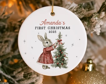 May include: A round white Christmas ornament with the text "Amanda's FIRST CHRISTMAS 2025". It features a bunny in a red dress decorating a Christmas tree. The ornament is suspended by a gold string, and is decorated with snowflakes.