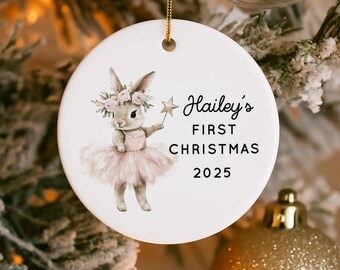 May include: A round white ceramic Christmas ornament featuring a bunny in a pink tutu and floral crown. The text reads "Hailey's FIRST CHRISTMAS 2025". A gold string is attached for hanging. The ornament is decorated with a gold star.