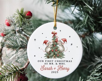 May include: A round white Christmas tree decoration with a gold hanging loop. It depicts two mice in Santa hats decorating a Christmas tree. The text reads "Our First Christmas as Mr. & Mrs. Sarah + Henry 2025."