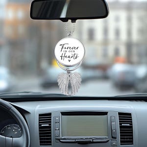 May include: A silver car charm with angel wings and a circular pendant. The pendant has the text "Forever in our Hearts" in black script. The charm hangs from a chain and is suspended in front of a car's dashboard.