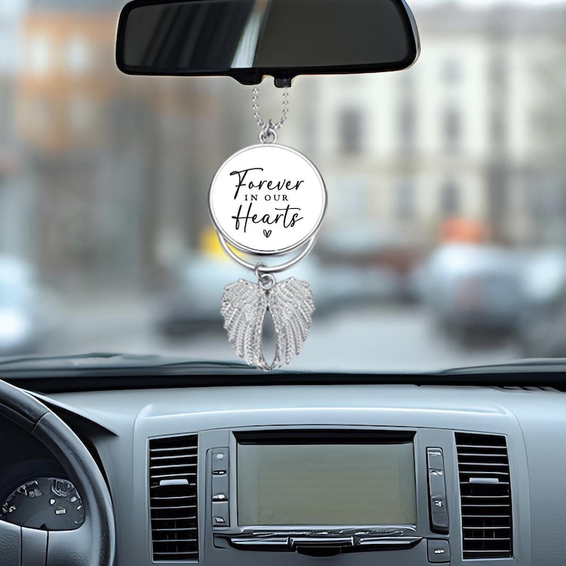 May include: A silver car charm with angel wings and a circular pendant. The pendant has the text "Forever in our Hearts" in black script. The charm hangs from a chain and is suspended in front of a car's dashboard.
