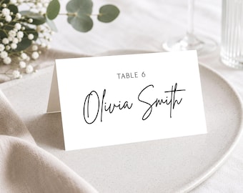 May include: A white folded paper place card featuring the printed text TABLE 6 and the name Olivia Smith in a black handwritten-style script font. The card is positioned upright on a neutral-coloured plate, displaying clear black typography.