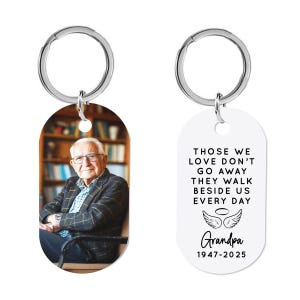 May include: A white oval keychain with a black and white photo of a man on one side and a black and white text message on the other side. The text reads "Those we love don't go away they walk beside us every day. Grandpa 1947-2025".
