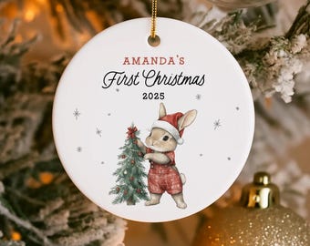 May include: A round white Christmas ornament with the text "AMANDA'S First Christmas 2025". It features a bunny in a Santa hat and red dungarees, decorating a Christmas tree. The ornament is suspended by a gold string, perfect for festive decor.