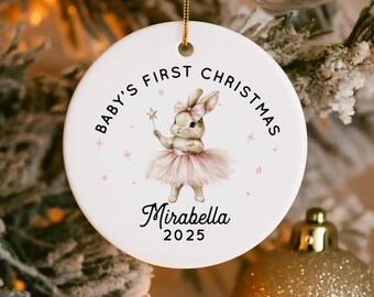 May include: A white ceramic Christmas bauble with a gold hanging loop. It features a bunny in a pink tutu holding a star wand. Text reads "Baby's First Christmas" and "Mirabella 2025". The ornament is hanging on a Christmas tree.