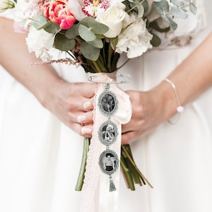 May include: A bridal bouquet charm with three oval-shaped photo frames, each with a black and white photo of a couple. The charm is attached to a white satin ribbon with a pink lace trim and a tassel.