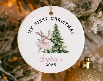 May include: Round white ceramic Christmas ornament with a gold hanging string. It features a bunny in a pink tutu, a Christmas tree, and the text "My First Christmas" and "Sophia's 2025".