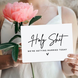 May include: White card with black calligraphy that reads "Holy Shit We're Getting Married Today" with a small heart symbol below the text. A person is holding the card in front of a pink peony.