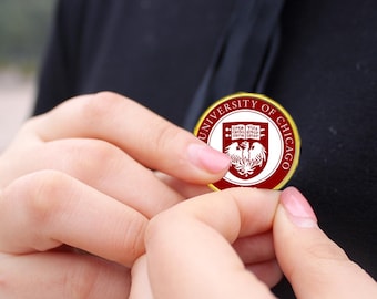 May include: A circular pin with a gold edge, displaying the words "UNIVERSITY OF CHICAGO". The pin features the university's emblem in red and white. The pin is held between two hands.