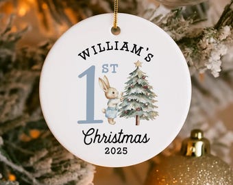 May include: Round white ceramic Christmas ornament with gold trim. It features the text "William's 1st Christmas 2025." A blue number 1 and a watercolour illustration of a bunny and Christmas tree are also on the ornament.