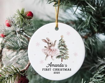 May include: Round white ceramic Christmas ornament featuring a bunny in a pink tutu and a Christmas tree illustration. The text reads "Amanda's FIRST CHRISTMAS 2025". The ornament hangs from a gold cord.