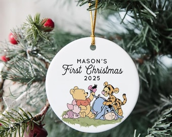 May include: A white, round Christmas ornament with the text "MASON'S First Christmas 2025". The ornament features illustrations of Winnie the Pooh, Piglet, Eeyore, and Tigger. A gold string is attached for hanging.