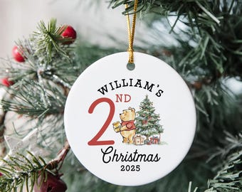 May include: Round white ceramic Christmas ornament with gold string. It features the text "William's 2nd Christmas 2025." A cartoon bear holds a honey pot next to a Christmas tree and wrapped gifts. The ornament hangs on a Christmas tree.