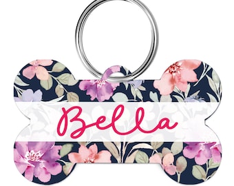 May include: A bone-shaped dog tag with a floral pattern in shades of pink, purple, and green on a navy blue background. The tag has a white band with the name "Bella" written in pink cursive.