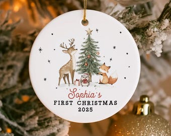 May include: A circular white Christmas ornament with a gold hanging string. It features a watercolour illustration of a deer, fox, Christmas tree, and a small animal. The text reads "Sophia's First Christmas 2025".