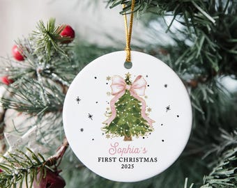 May include: A white, circular Christmas ornament featuring a watercolour illustration of a Christmas tree adorned with a pink bow and ribbon. The text reads "Sophia's First Christmas 2025". It hangs from a gold string, perfect for festive decor.
