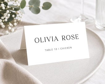 May include: A white folded place card with black serif typography reading OLIVIA ROSE and TABLE 10 | CHICKEN. The card is positioned on a neutral-toned plate, featuring a clean, minimalist design suitable for formal event seating arrangements.