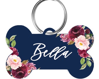 May include: A navy blue dog tag in a bone shape, featuring the name "Bella" in white script. The tag is adorned with floral watercolor designs in shades of burgundy, pink, and green, and includes a silver metal ring for attachment.