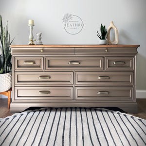 May include: A light brown dresser with eight drawers and brass hardware. The dresser has a wood top and sits on a striped rug.
