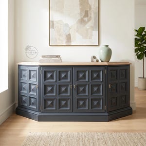 May include: A dark gray sideboard featuring a light wood top and four cabinet doors with a raised square geometric pattern. The unit has a multi-faceted front design with small metallic hinges and handles, providing storage space for home organization.
